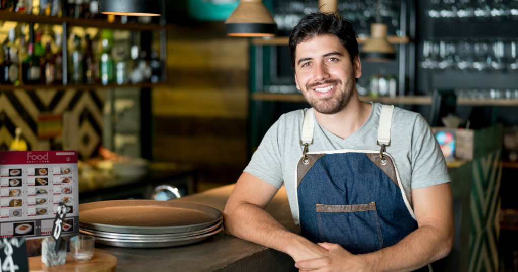 Smiling restaurant owner in apron at a busy bar, showcasing business growth and cash flow solutions for seasonal success.