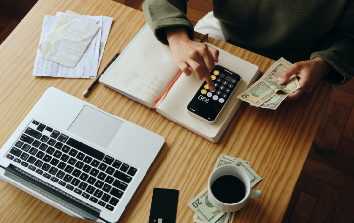 Person calculating finances with a calculator, laptop, cash, and notebook, emphasizing fiscal literacy and money management skills.