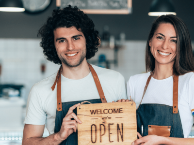 Smiling baristas holding a 'Welcome, we're OPEN' sign, showcasing excellent customer service and connection strategies.