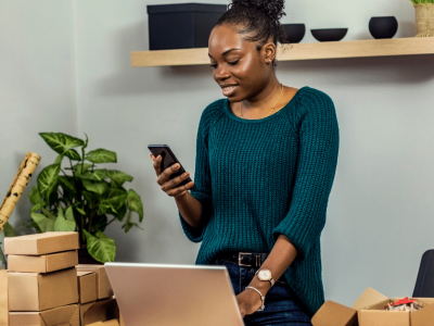 Woman in a green sweater using a smartphone in a small business setting with packages and a laptop, exploring bad credit loan options.
