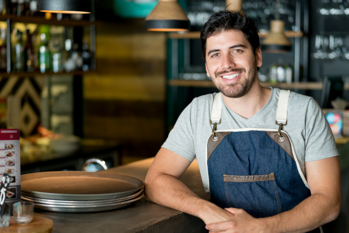 Smiling restaurant owner in apron at a busy bar, showcasing business growth and cash flow solutions for seasonal success.