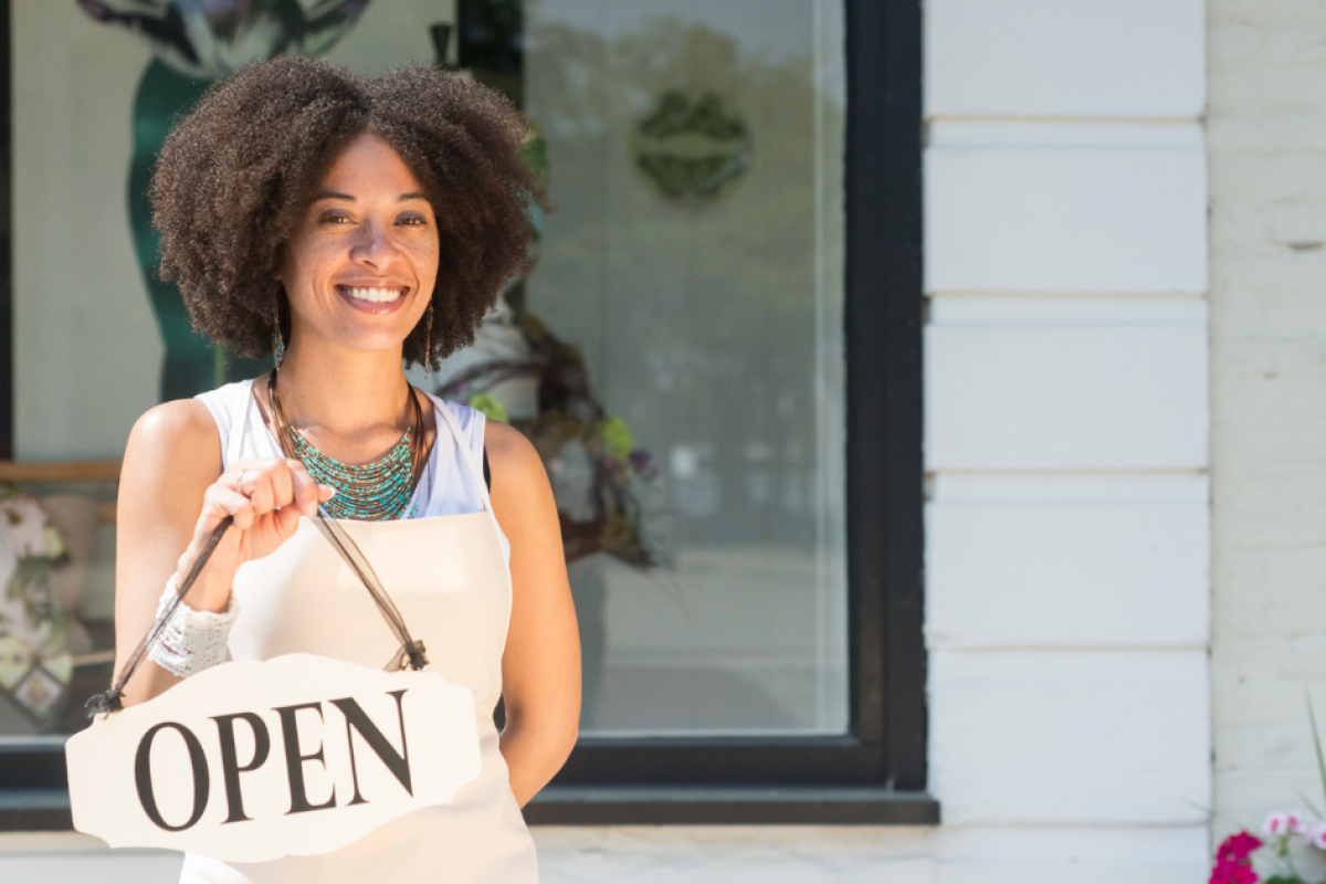 Smiling woman holding an 'OPEN' sign outside her business, representing women entrepreneurs and unsecured credit options.