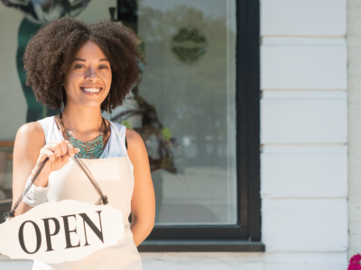 Smiling woman holding an 'OPEN' sign outside her business, representing women entrepreneurs and unsecured credit options.