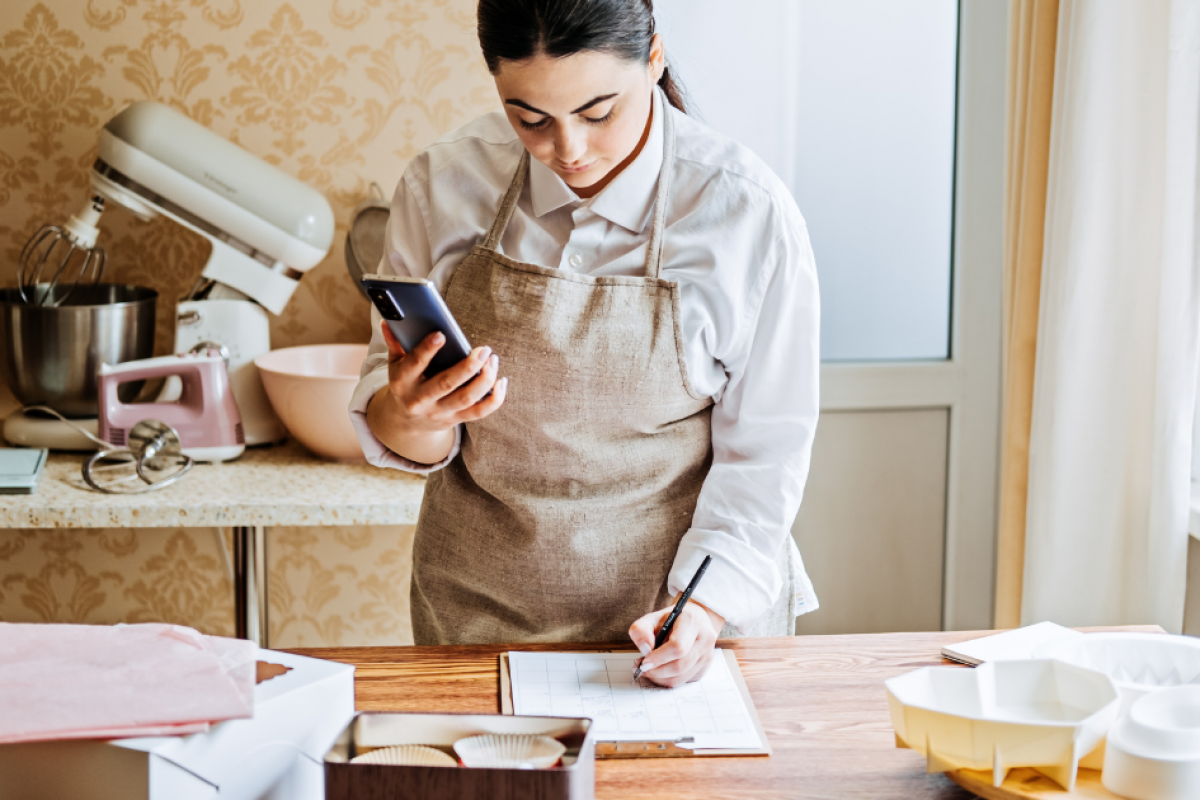 Woman in an apron using a phone while writing notes in a kitchen, preparing for a baking project.