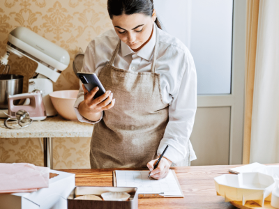Woman in an apron using a phone while writing notes in a kitchen, preparing for a baking project.