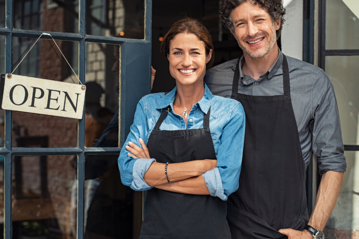 Smiling grocery business owners standing by an open sign, representing bad credit business loans for growth and cash flow.