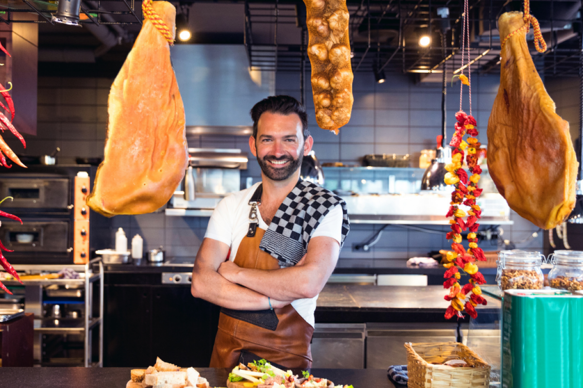 Chef in a restaurant kitchen with hanging meats and spices, showcasing effective business budget strategies for culinary success.