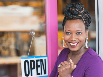 Smiling business owner in front of an open sign, representing small business empowerment and cash advance solutions.
