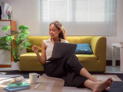 Young businesswoman sitting on the floor in front of her sofa is using a laptop and a credit card to shop online from home, enjoying the comfort and convenience of e-commerce