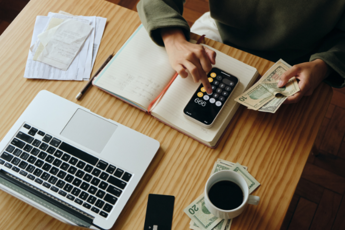 Person calculating finances with a calculator, laptop, cash, and notebook, emphasizing fiscal literacy and money management skills.